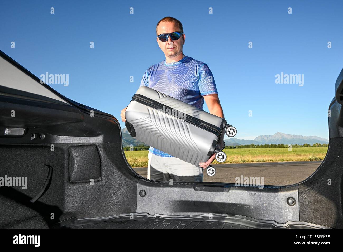 Mann, der Koffer in den Kofferraum steckt, bereit für Roadtrip-Abenteuer in landschaftlich reizvoller Lage. Stockfoto