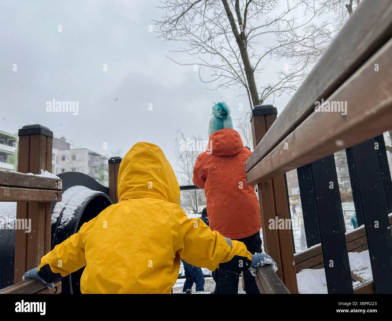 Kinder in warmer Winterkleidung spielen auf einem schneebedeckten Spielplatz. Die leuchtend gelben und orangefarbenen Jacken stehen im Kontrast zur verschneiten Landschaft cr Stockfoto