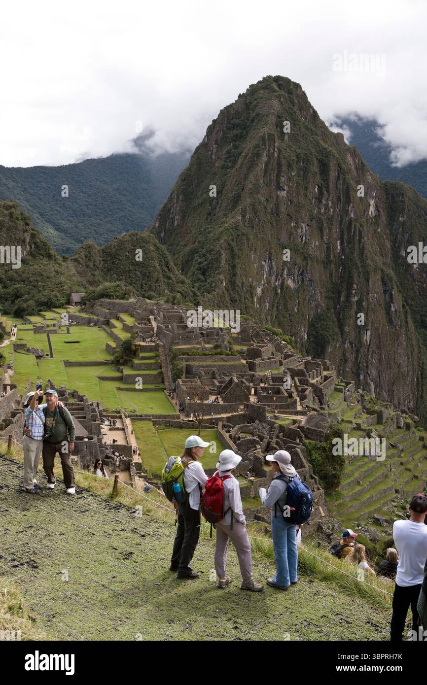 Zwei weibliche Touristen hören ihrem Reiseleiter zu, während sie Machu Picchu (mit dem Berg Huayna Picchu dahinter) in Peru überblicken. Stockfoto