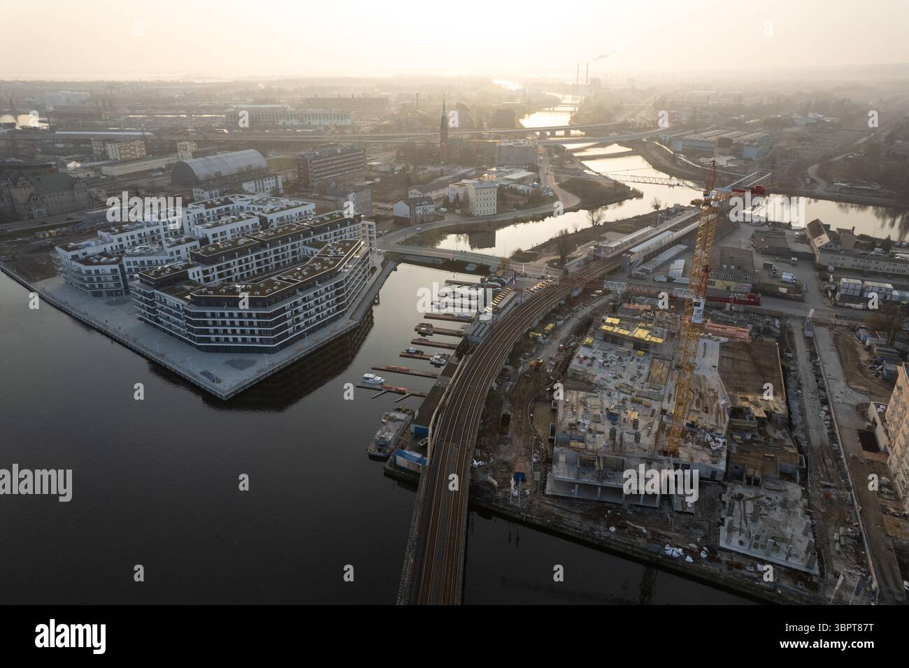 Drohnenblick auf eine Stadtlandschaft bei Sonnenuntergang oder Sonnenaufgang. Das Bild zeigt einen großen, modernen Wohnkomplex am Flussufer neben einem großen Konstru Stockfoto
