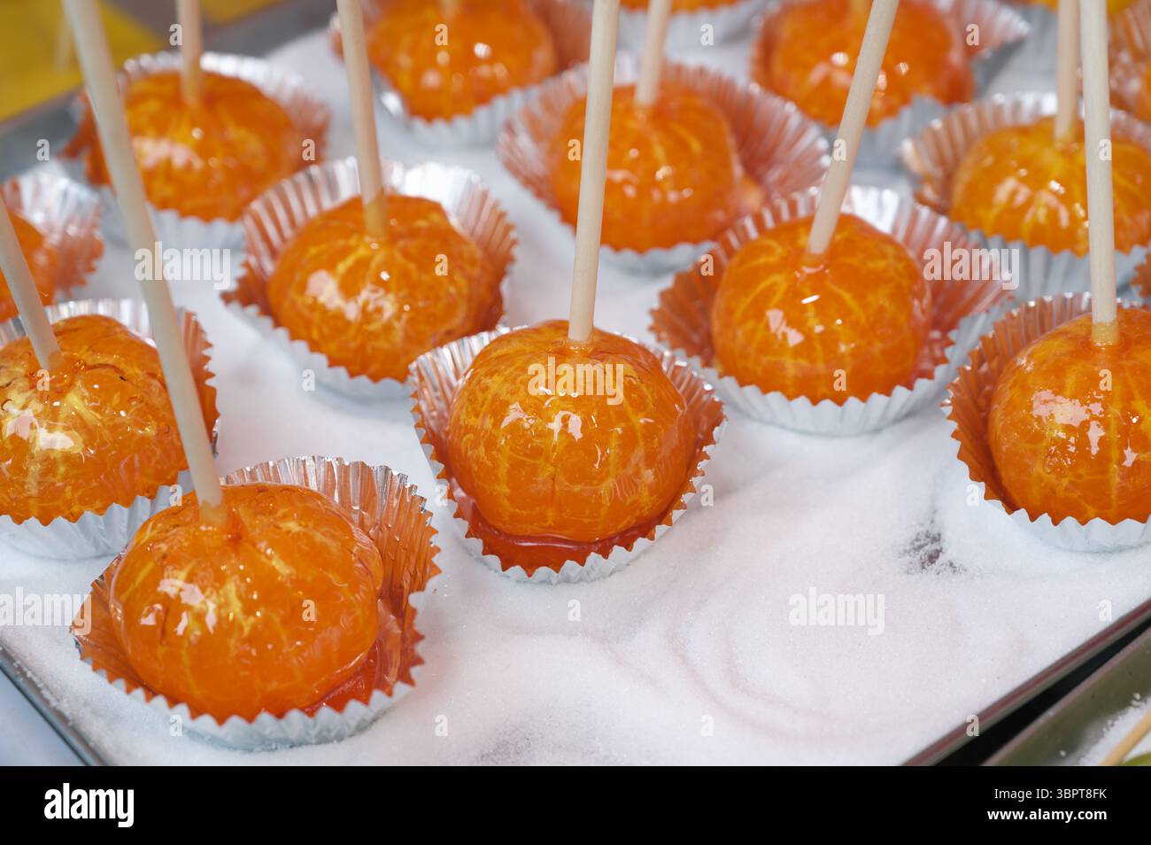 Glasierte kandierte Orangen auf Sticks auf einem Tokioter Lebensmittelmarkt, ausgestellt in glänzenden Folientassen über einem Bett mit Zucker Stockfoto