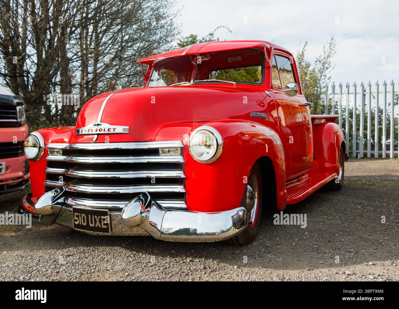 Roter Chevrolet 3100 Pickup-Truck mit Chromakzenten, der auf einer unbefestigten Straße neben einem weißen Streifenzaun und Bäumen geparkt ist Stockfoto