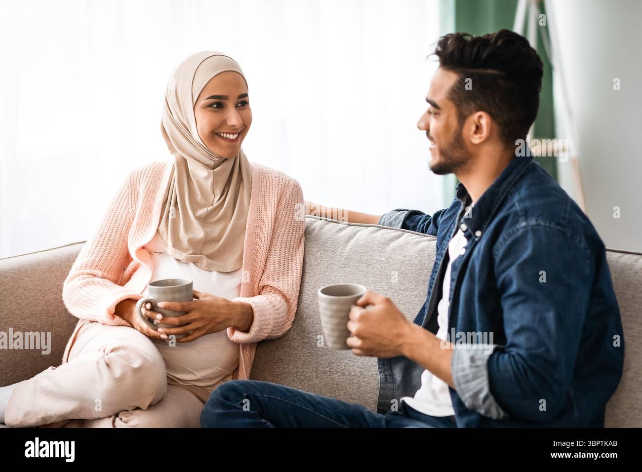 Fröhliches, Schwanger-Muslimisches Paar, Das Sich Zu Hause Ausruhte, Kaffee Trinkt Und Sich Unterhielt Stockfoto