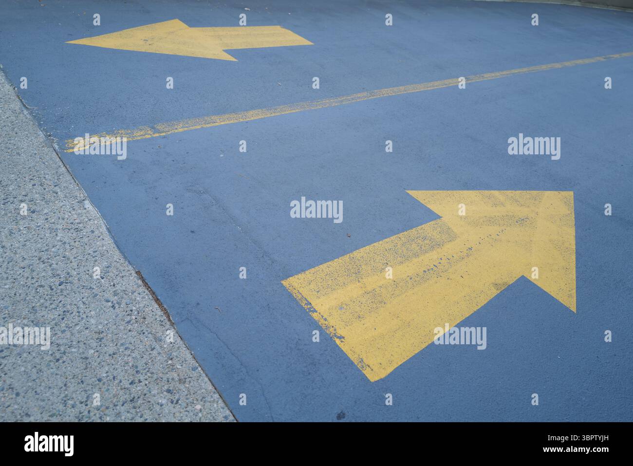 Zwei kräftige gelbe Pfeile auf blauem Asphaltboden, die in entgegengesetzte Richtungen zeigen, symbolisieren Wahl oder Entscheidung. Stockfoto