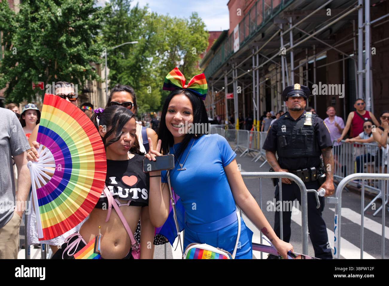 Zwei junge Frauen, eine mit Regenbogenschleife und blauem Hemd, die andere ein schwarzes Oberteil, posieren bei einer Pride-Parade. Ein Regenbogen-Handventilator wird von einem der Frauen gehalten Stockfoto
