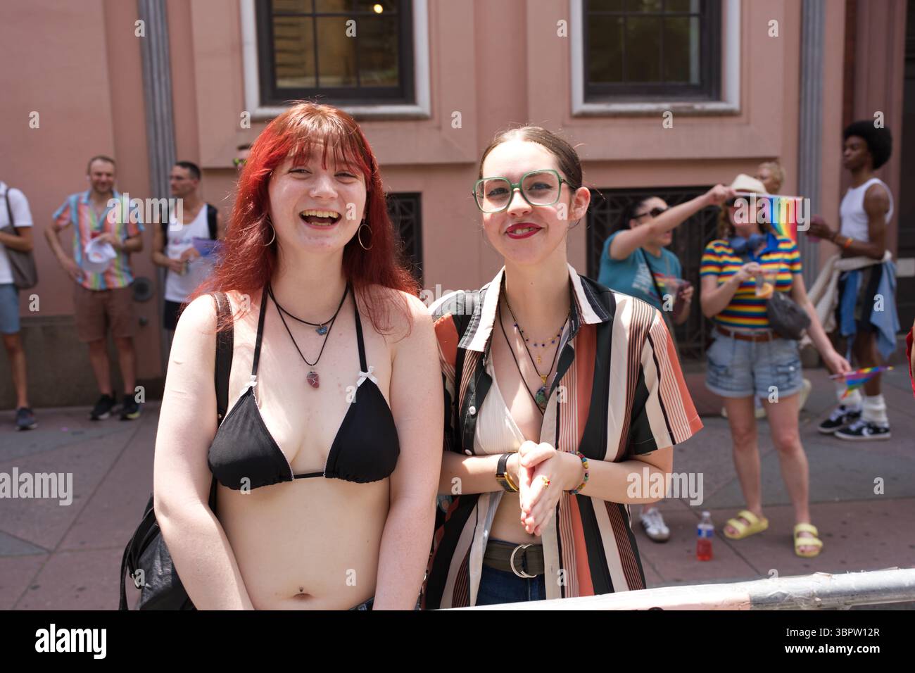 Zwei junge Frauen, eine mit rotem Haar und schwarzem Bikinioberteil, die andere mit braunem Haar und gestreiftem Hemd, stehen draußen lächelnd und unscharf Stockfoto