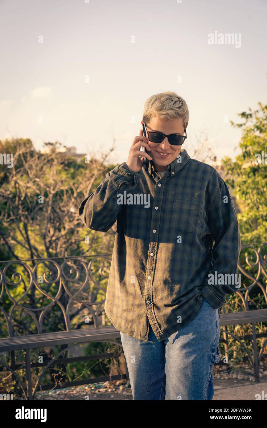 Ein lächelnder junger Mensch mit einem modernen, androgynen Aussehen spricht draußen auf einem Smartphone. Konzept des zeitgenössischen Lebensstils, der Kommunikation, der Jugendkultur Stockfoto