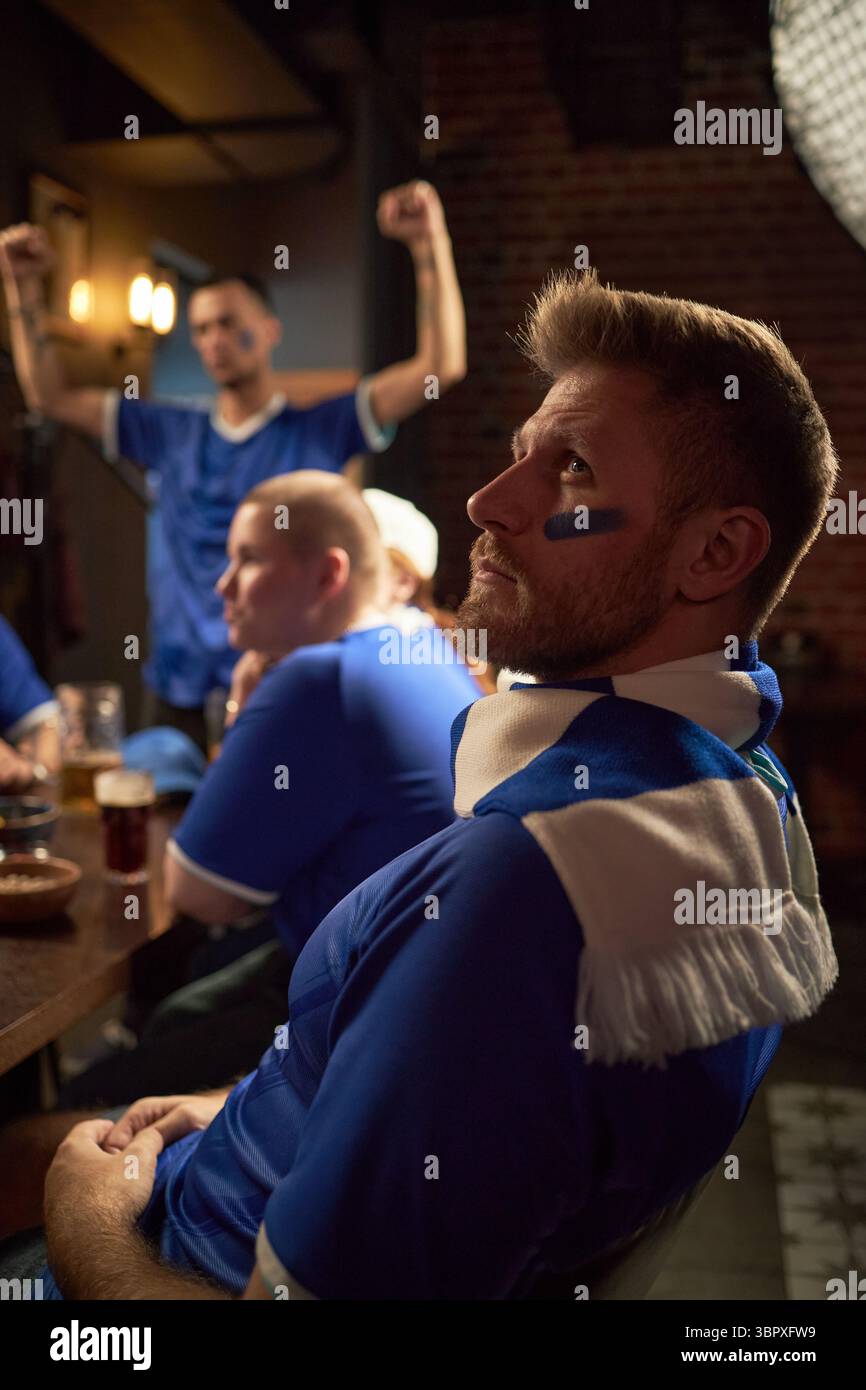 Kaukasischer junger männlicher Fan mit gemaltem Gesicht, der am Tisch sitzt und Fußballspiele mit einer Gruppe multiethnischer junger Freunde in der Sportbar beobachtet. Hintergrund zeigt den Mann, der feiert Stockfoto