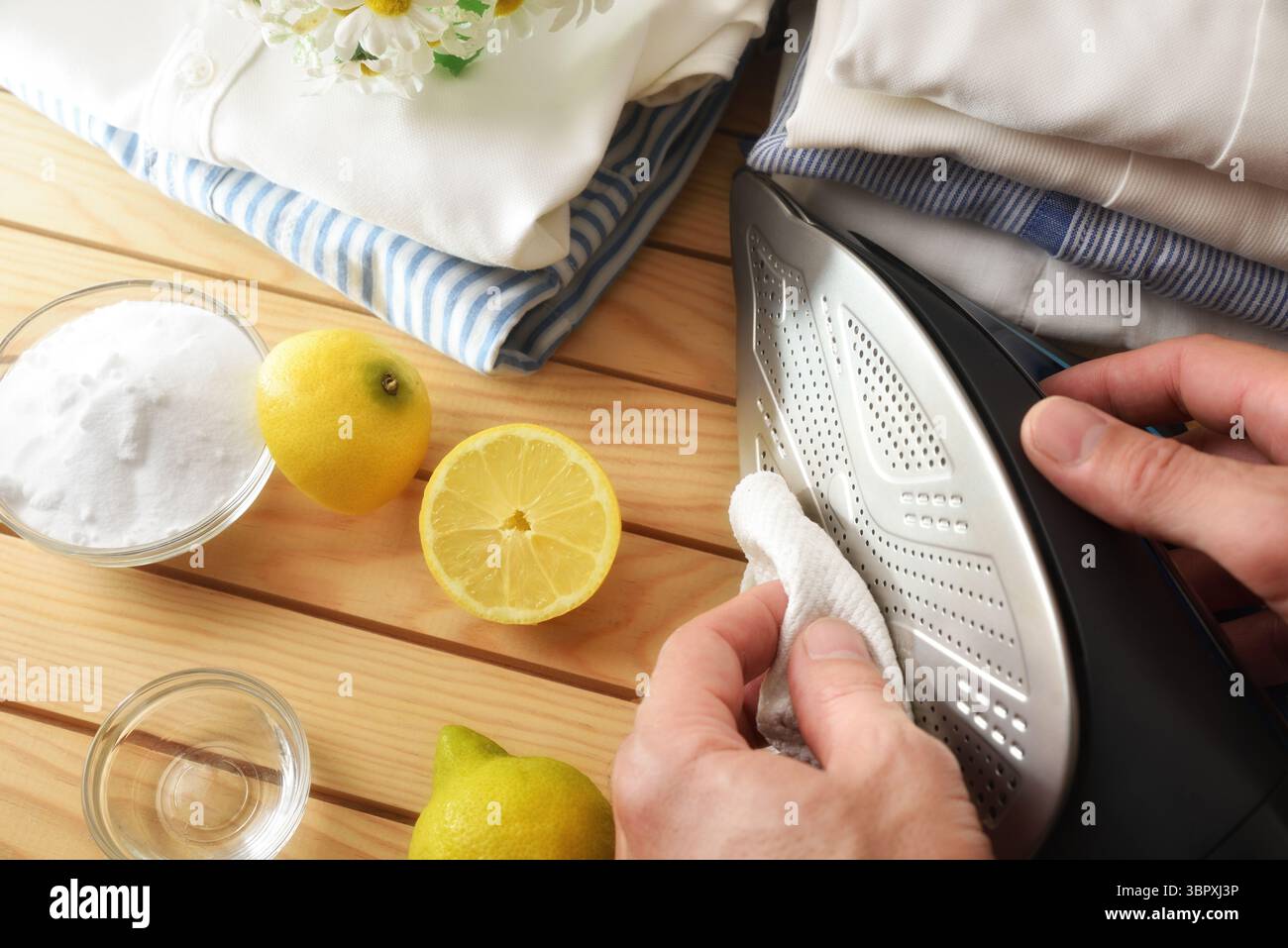 Person, die die Bügelsohle eines Dampfbügeleisens mit einem Tuch und selbstgemachten Naturprodukten auf einem Holztisch und gebügelten Hemden im Hintergrund reinigt. Top VI Stockfoto