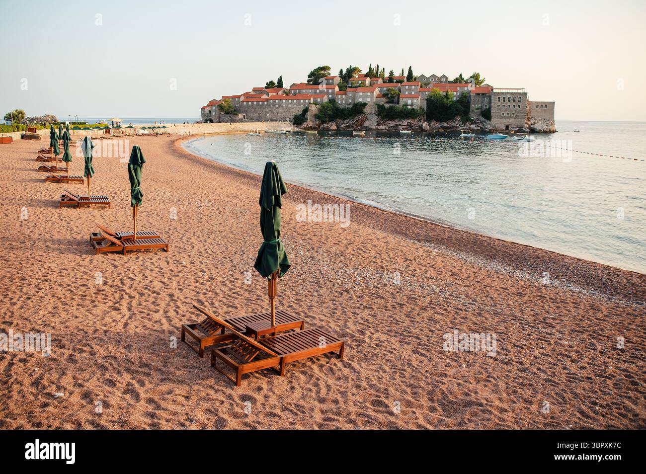 Leerer Strand mit Holzliegen und geschlossenen Sonnenschirmen mit Blick auf eine historische Inselstadt. Stockfoto