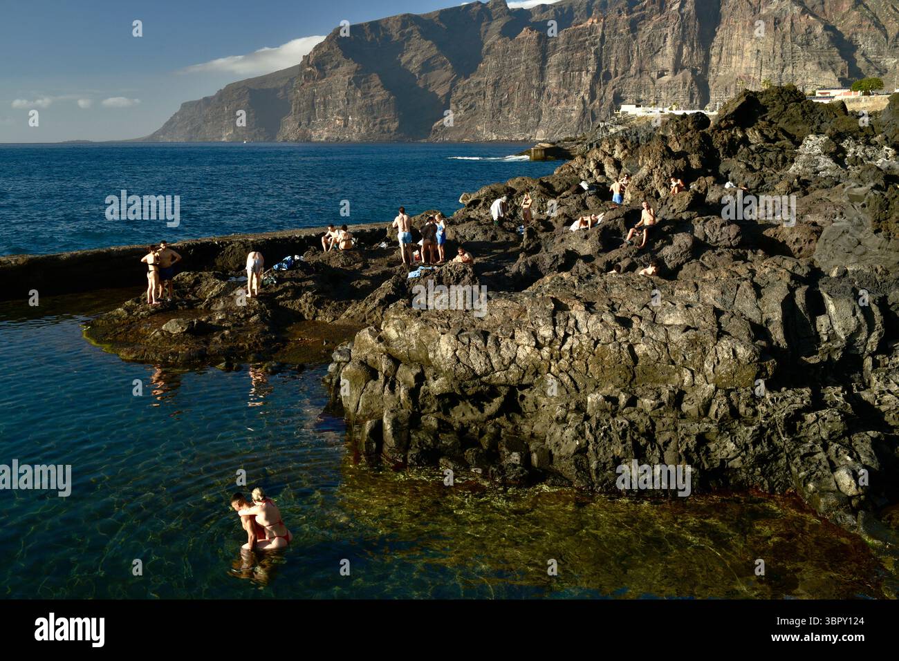 Menschen schwimmen in natürlichen Küstenbecken mit Los Gigantes Bergen und Klippen in der Ferne, Charco de Isla Cangrejo, Teneriffa, Kanarische Inseln, Spanien Stockfoto