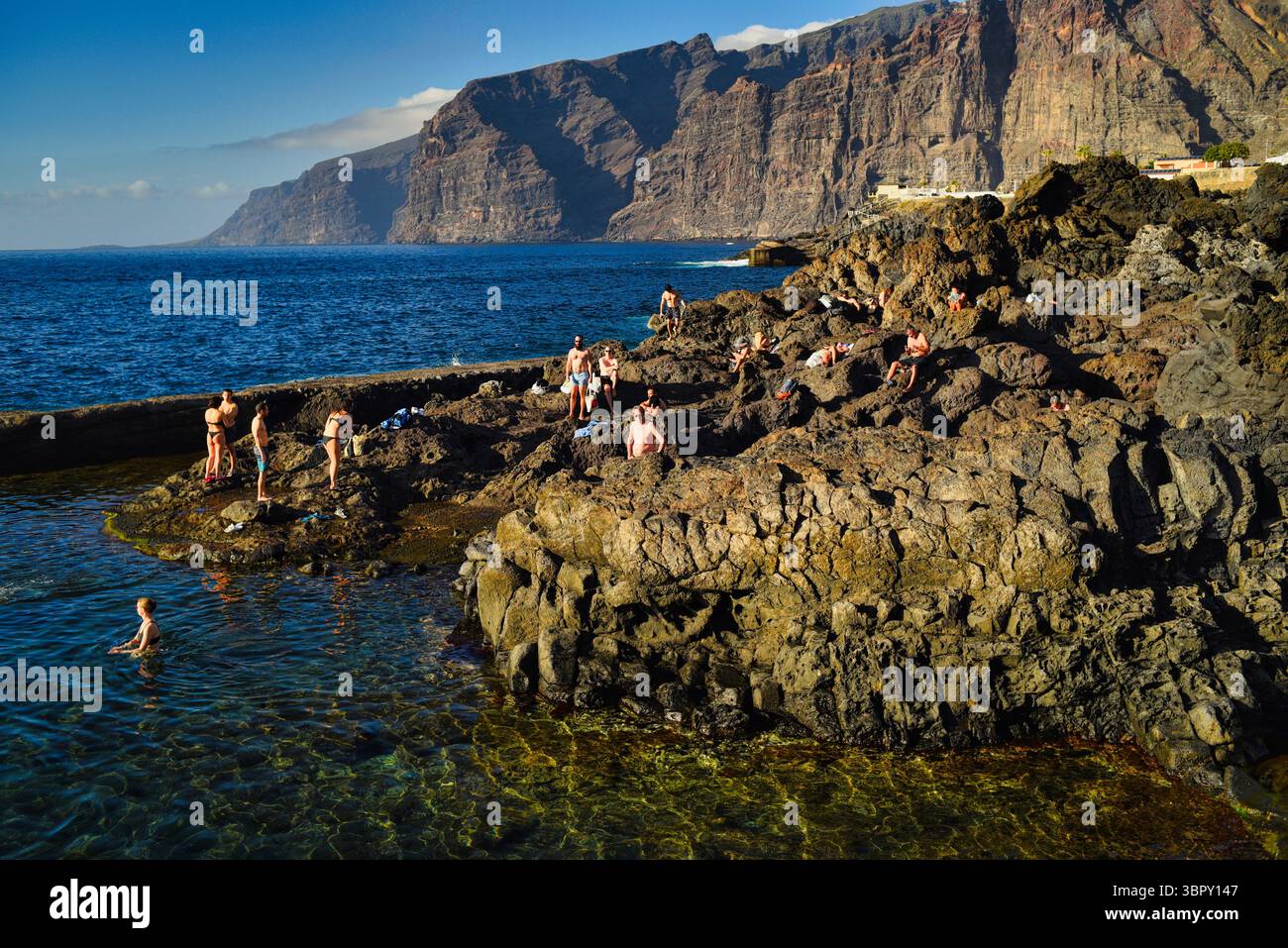 Menschen schwimmen in natürlichen Küstenbecken mit Los Gigantes Bergen und Klippen in der Ferne, Charco de Isla Cangrejo, Teneriffa, Kanarische Inseln, Spanien Stockfoto