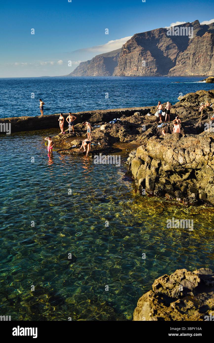 Menschen schwimmen in natürlichen Küstenbecken mit Los Gigantes Bergen und Klippen in der Ferne, Charco de Isla Cangrejo, Teneriffa, Kanarische Inseln, Spanien Stockfoto