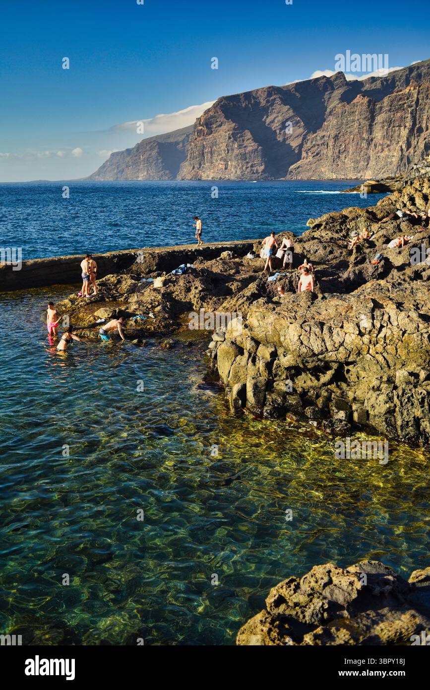 Menschen schwimmen in natürlichen Küstenbecken mit Los Gigantes Bergen und Klippen in der Ferne, Charco de Isla Cangrejo, Teneriffa, Kanarische Inseln, Spanien Stockfoto