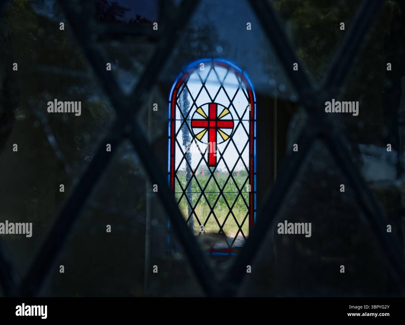 Sanftes Sonnenlicht strömt durch das rote Buntglaskreuz und strahlt warmes, ätherisches Licht im Inneren der historischen Kirche aus Stockfoto