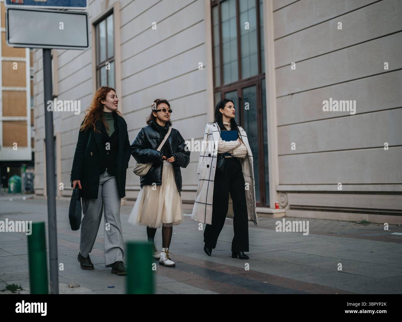 Drei stilvolle Frauen in modischen Mänteln interagieren warm, während sie an einem frischen Herbsttag eine Straße entlang laufen. Stockfoto