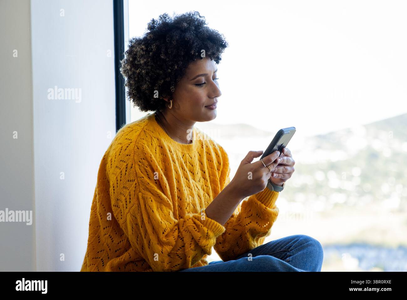 Afroamerikanische Frau in Senfpullover, die am Fenstersitz sitzt und den Bildschirm des Smartphones hält. Lässig, modern, gemütlich, Lifestyle, natürlich, städtisch, Stockfoto