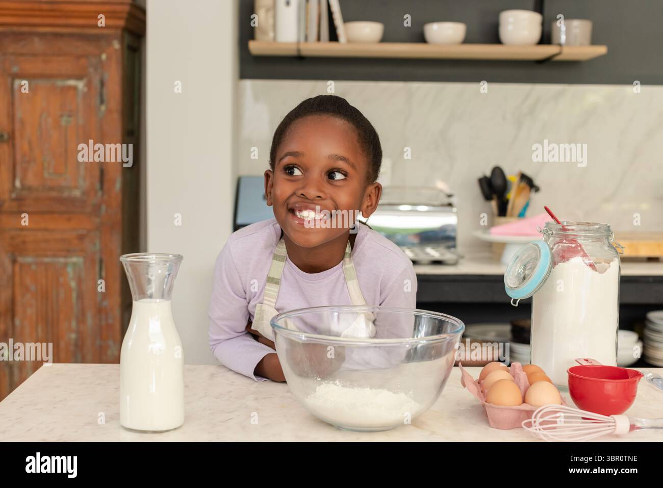 Lächelndes afroamerikanisches Mädchen in Schürze, Mehl und Milch in einer Glasschale in der Küche zu Hause. Kinder, kulinarisch, rustikal, Hausmannskost, fröhlich, Domest Stockfoto