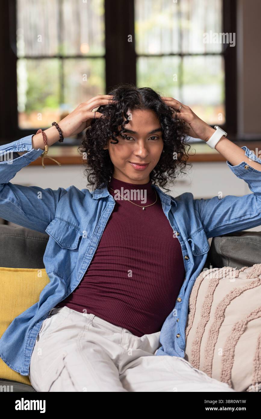 Nicht binärer Erwachsener, der auf dem Sofa sitzt und lockiges Haar im Wohnzimmer mit Smartwatch anpasst. Modern, lässig, innen, Komfort, Wellness, Diversität, lifestyl Stockfoto