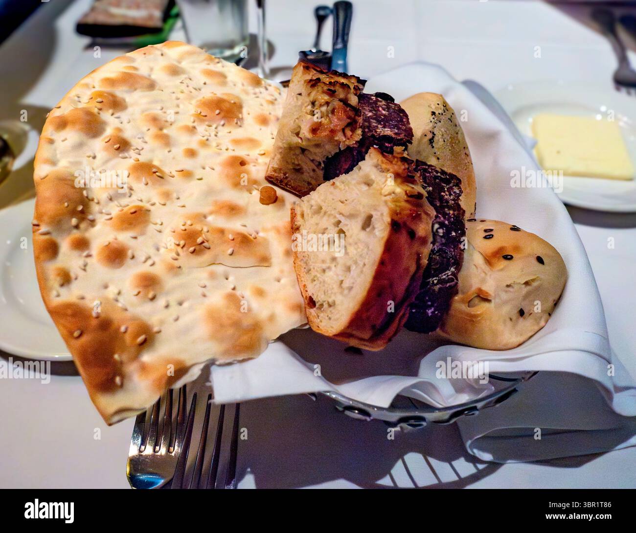 Ein Brotkorb liegt auf einer weißen Tischdecke, mit einem großen Sesamfladenbrot und verschiedenen Brötchen, einige mit Kräutern oder Füllungen. Ein Klecks Butter sitzt n Stockfoto