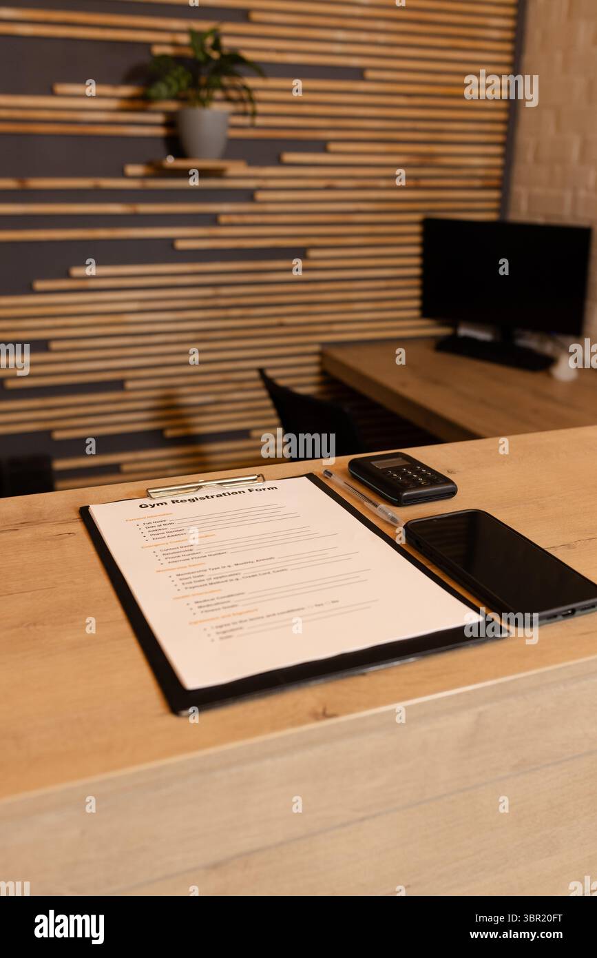 Clipboard mit dem Anmeldeformular für das Fitnessstudio, das auf dem Rezeptionstresen zwischen Stift, Pflanze und Lamellenwand sitzt. Fitness, Professionalität, modern, minimalistisch, Technologie Stockfoto