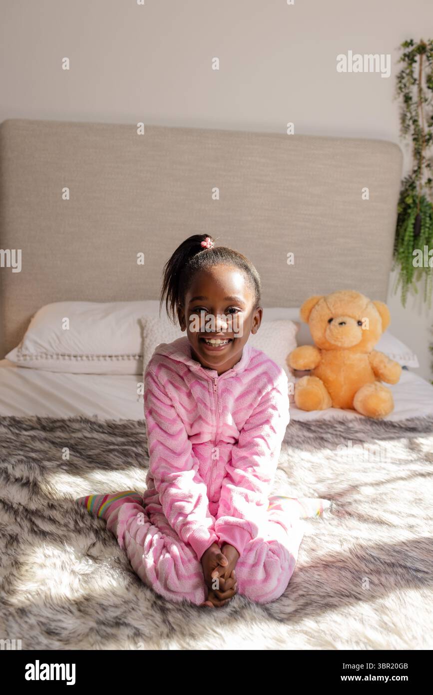 Afroamerikanisches Mädchen mit Pyjama-Einteiler, das auf dem Bett sitzt und einen orangen Teddybären hält. Gemütlich, Kindheit, Entspannung, Zuhause, Komfort, Unschuld Stockfoto