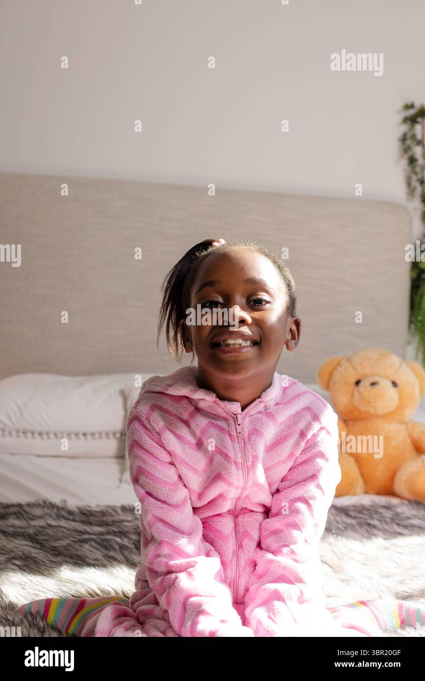 Afroamerikanerin, die im Schlafzimmer auf dem Bett sitzt und neben dem orangen Teddybär rosa, flauschige Einteiler trägt. Gemütlich, Komfort, Kindheit, Entspannung, Zuhause, Spielfu Stockfoto
