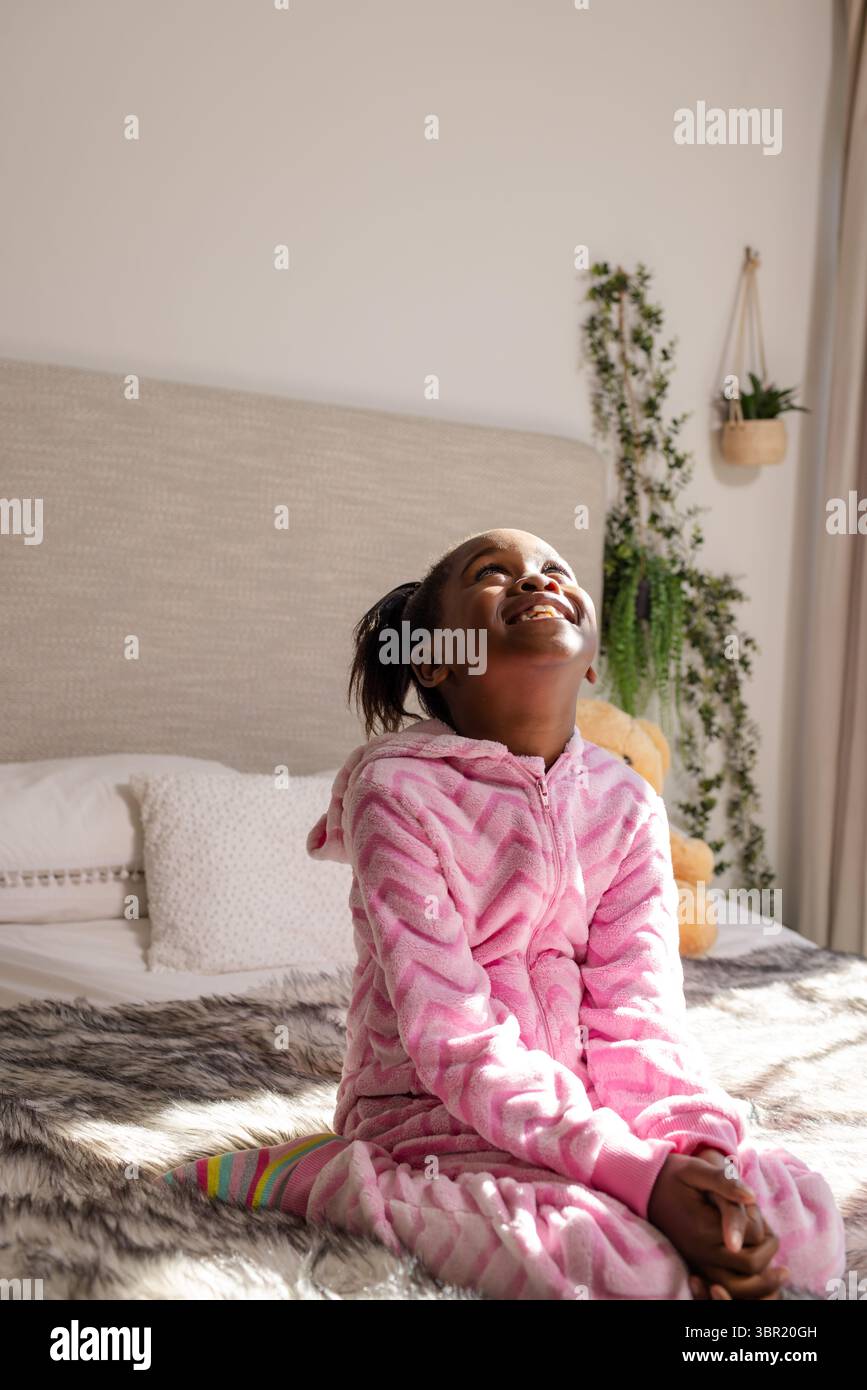 Afroamerikanisches Mädchen, das auf dem Bett sitzt, trägt rosa Einteiler und lächelt unter Sonnenlicht mit Teddybär. Verspielt, fröhlich, gemütlich, warm, jugendlich, fröhlich, Komm Stockfoto