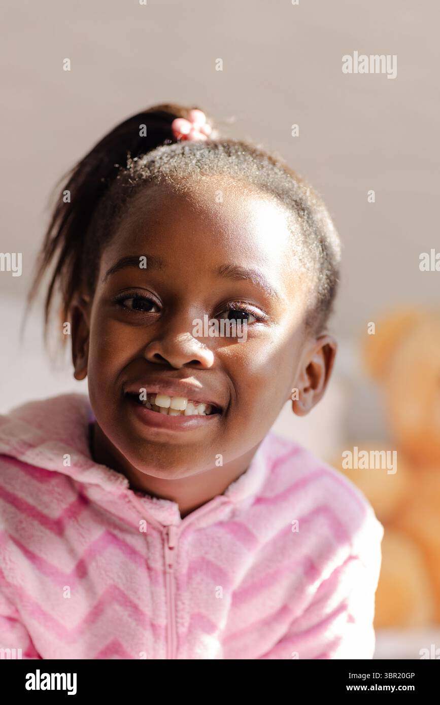 Afroamerikanisches Mädchen, das zu Hause sitzt und einen rosafarbenen Hoodie trägt, lächelt vor der Kamera mit einem Teddybären in der Nähe. Kinder, Kindheit, Trost, Unschuld, Wärme, r Stockfoto
