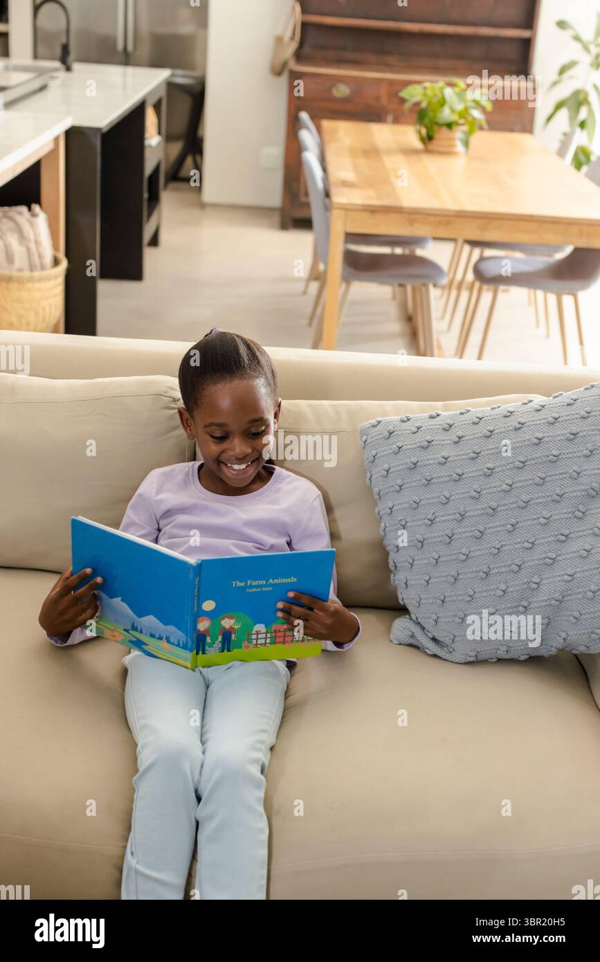 Afroamerikanisches Mädchen liest buntes Bilderbuch auf beigefarbenem Sofa im Wohnzimmer in der Nähe von Strickkissen. Kind, lesen, Zuhause, gemütlich, innen, Familie, entspannen Stockfoto