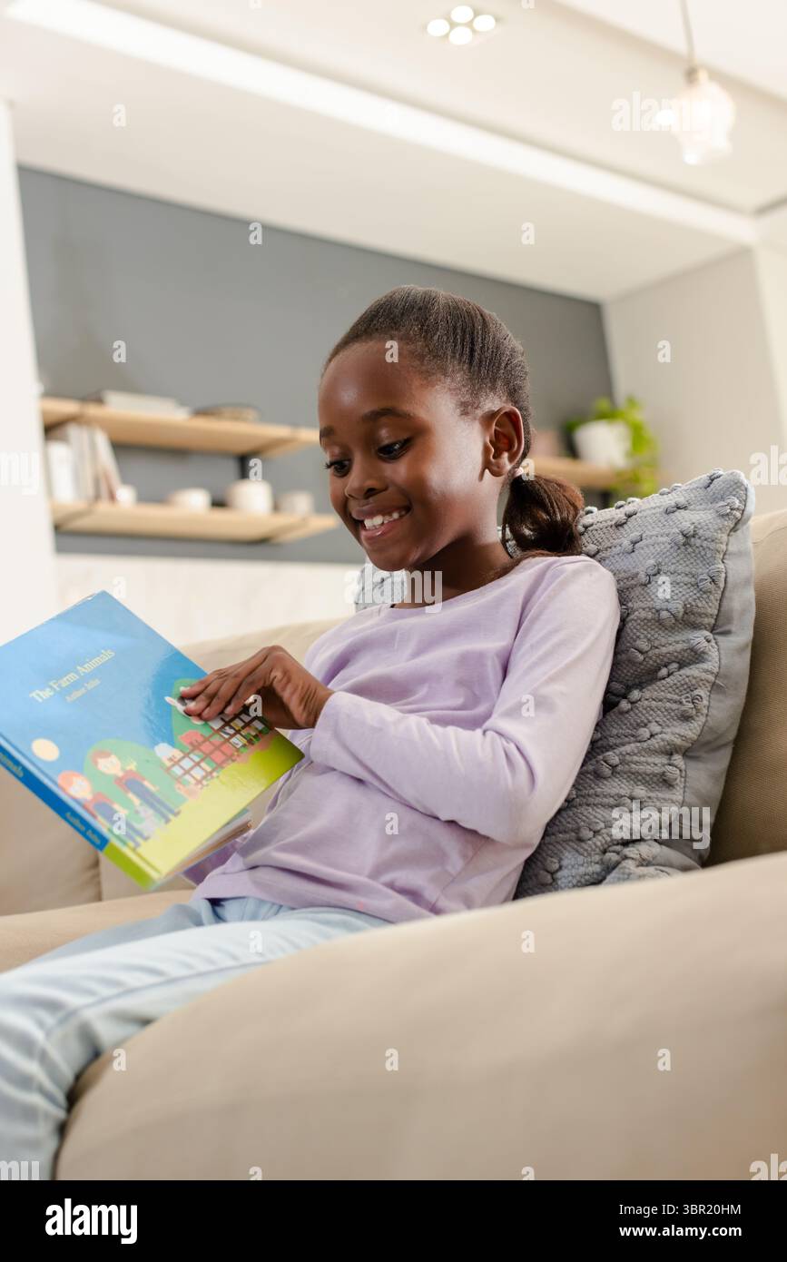 Afroamerikanisches Kind sitzt auf beigefarbenem Sofa im modernen Wohnzimmer und liest buntes Kinderbuch. Kind, lesen, Dekor, Inneneinrichtung, Bildung, Leisu Stockfoto
