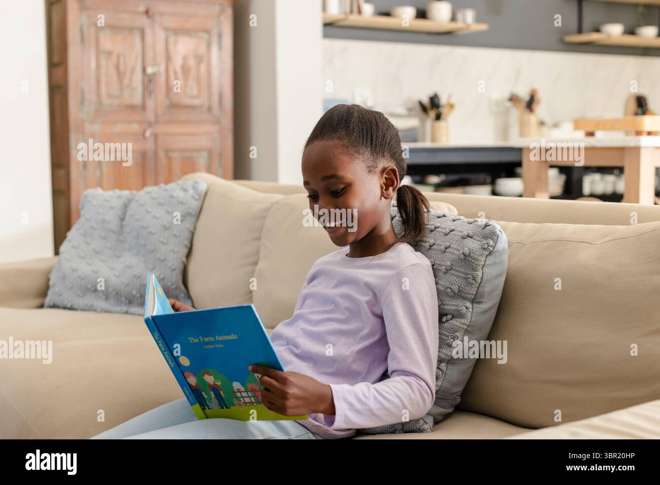 Afroamerikanisches Kind liest Kinderbuch auf beigefarbenem Sofa in der Nähe der Kücheninsel, Kopierraum. Gemütlich, einheimisch, bunt, lehrreich, Möbel, lifest Stockfoto