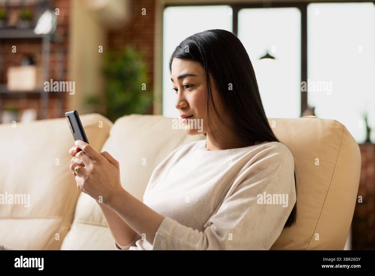 Asiatische Unternehmerin, die zu Hause auf der Couch sitzt, ihr Handy hält und soziale Medien überprüft. Externe Mitarbeiterin, die während der Arbeit von zu Hause aus eine Pause vom Laptop macht. Stockfoto