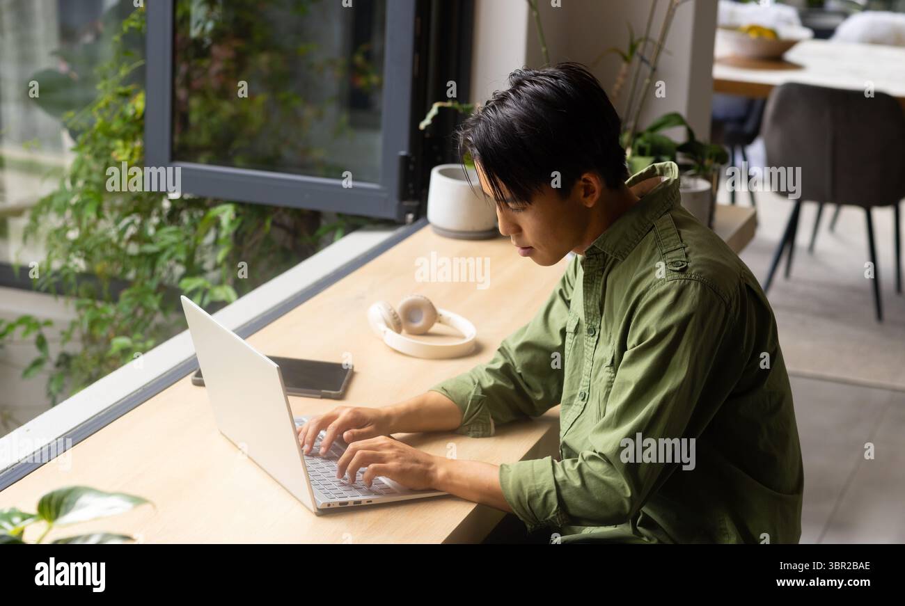 Mann tippt auf Laptop auf hölzerner Arbeitsplatte neben geöffnetem Fenster mit Smartphone-Kopfhörern und Pflanzen. Gemütlich, Produktivität, modern, Arbeitsbereich, Natur, ser Stockfoto