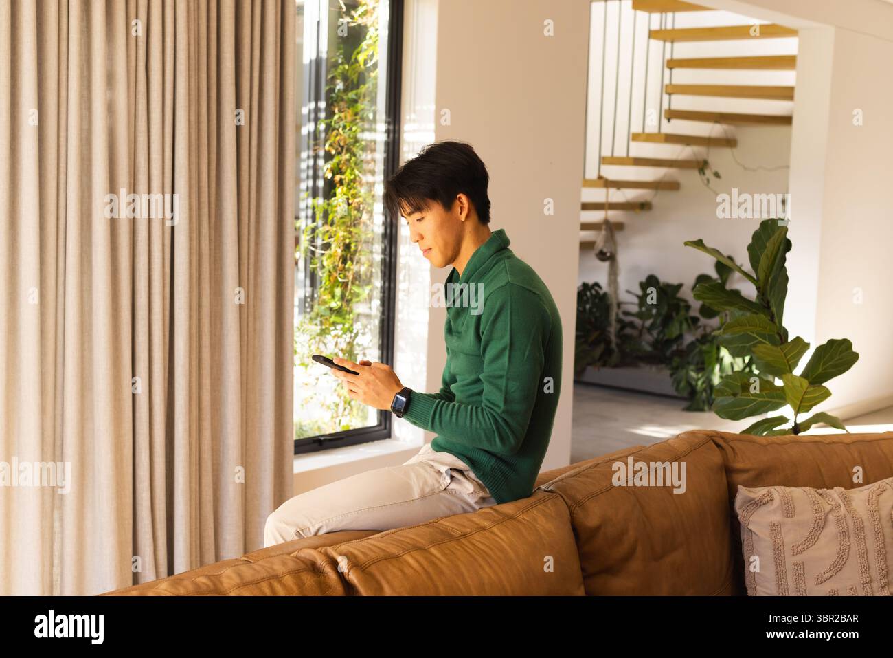 Erwachsener asiatischer Mann sitzt auf Couch mit beigefarbenen Vorhängen im Wohnzimmer mit Smartphone und Smartwatch. Modern, Interieur, Technologie, Lifestyle, Freizeit, Co Stockfoto