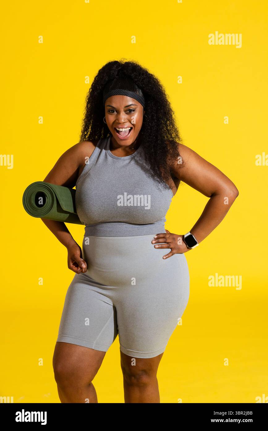 Afroamerikanerin, die im Studio steht und Sportkleidung trägt, mit gerollter Yogamatte und Smartwatch Stockfoto