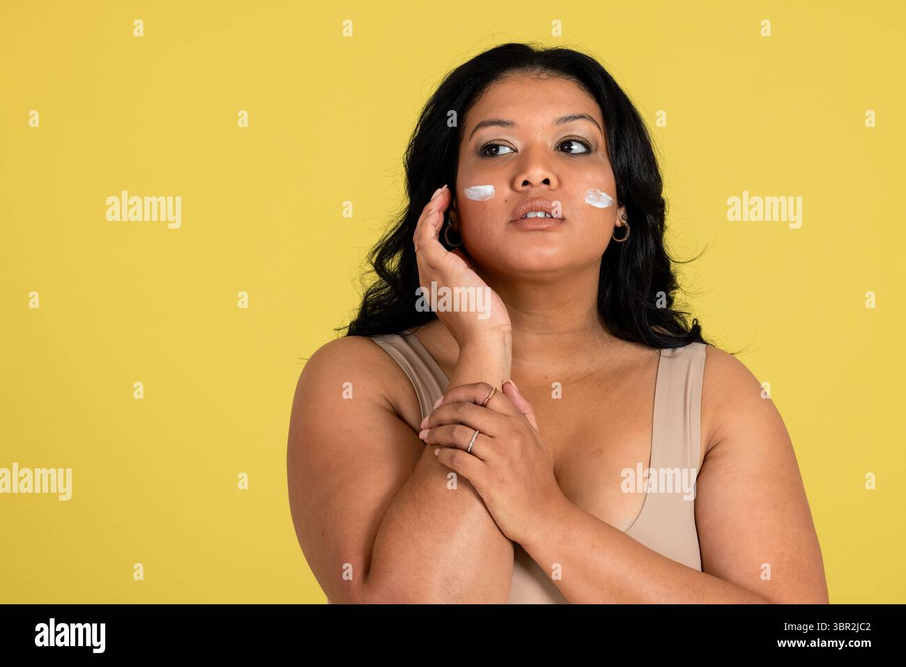 Asiatische Frau im Studio mit gelbem Hintergrund, die Hautpflegecreme auf die Wangen aufträgt Stockfoto
