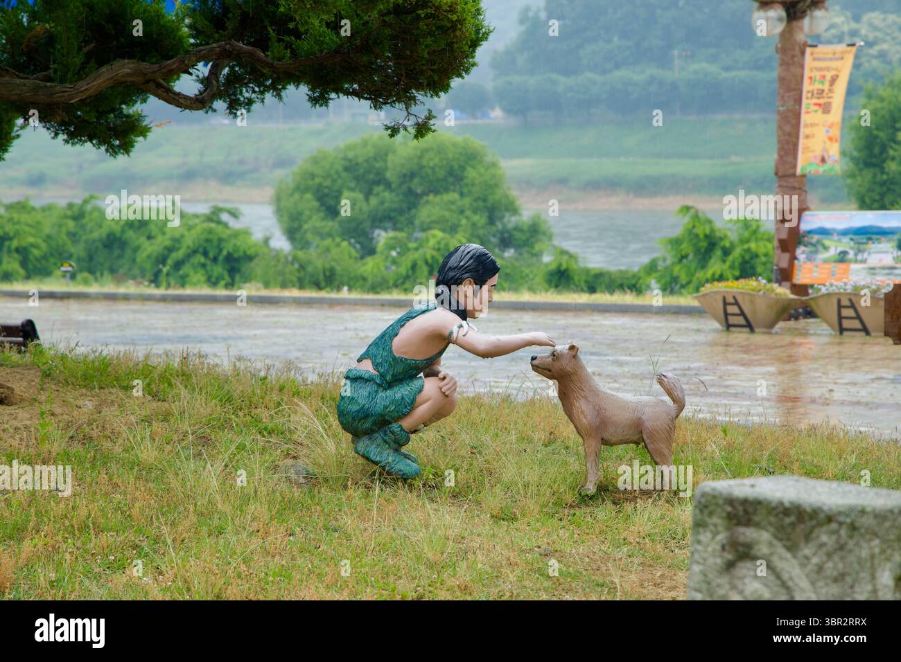 Gongju City, Südkorea - 13. Juni 2025: Eine Statue eines prähistorischen Kindes, das sanft einen Hund streichelt, steht auf dem grasbewachsenen Hügel in der Nähe des Geum River bei Seokj Stockfoto