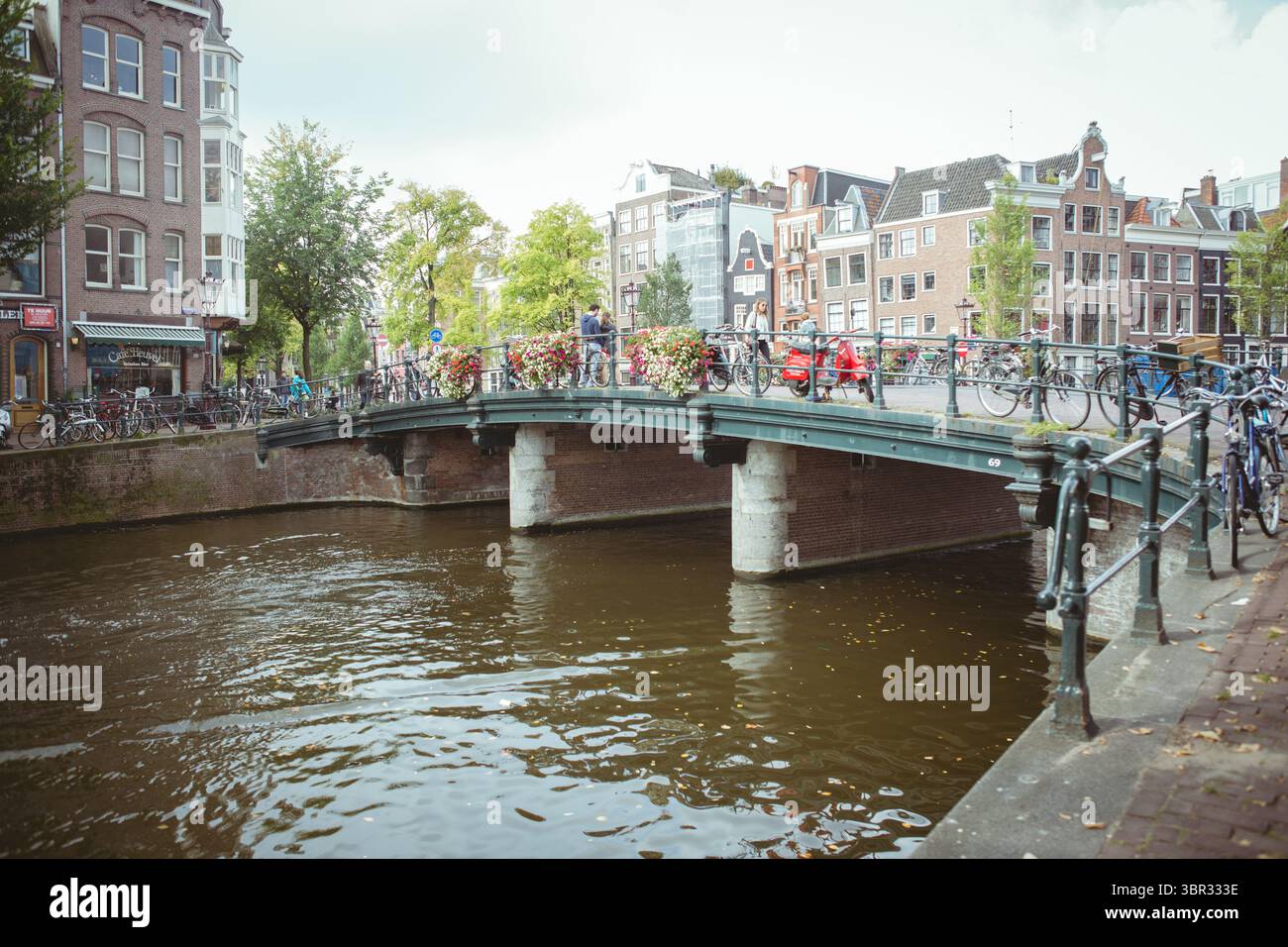 Das flache Design zeigt eine Fußgängerbrücke aus grünem Metall, die über einen braunen Kanal mit Blumenkörben verläuft Stockfoto