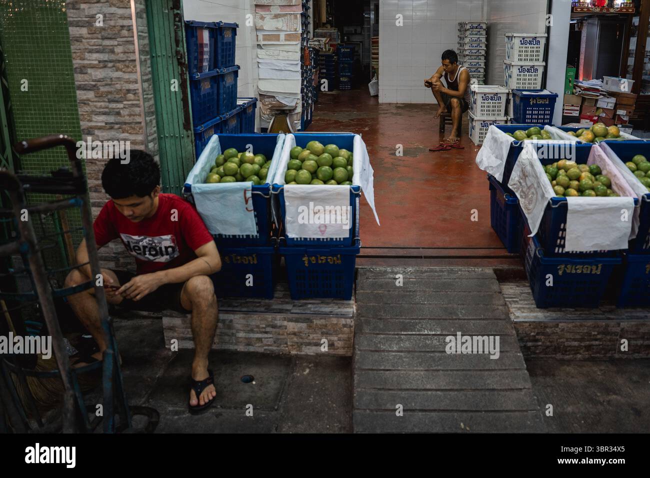 Bangkok, Thailand. Juli 2025. Zwei Männer spielen auf ihren Telefonen, während sie auf dem Markt neben Obstkisten sitzen. Jeden Morgen laufen buddhistische Mönche in Bangkok durch die Straßen, um Almosen, Essen und Blumen von den Laien zu erhalten. In der Nähe, auf dem Yodpiman Blumenmarkt, werden Blumen für diese täglichen Rituale zubereitet und verkauft, die in der buddhistischen Tradition Theravada verwurzelt sind. (Foto von Ploy Phutpheng/SOPA Images/SIPA USA) Credit: SIPA USA/Alamy Live News Stockfoto