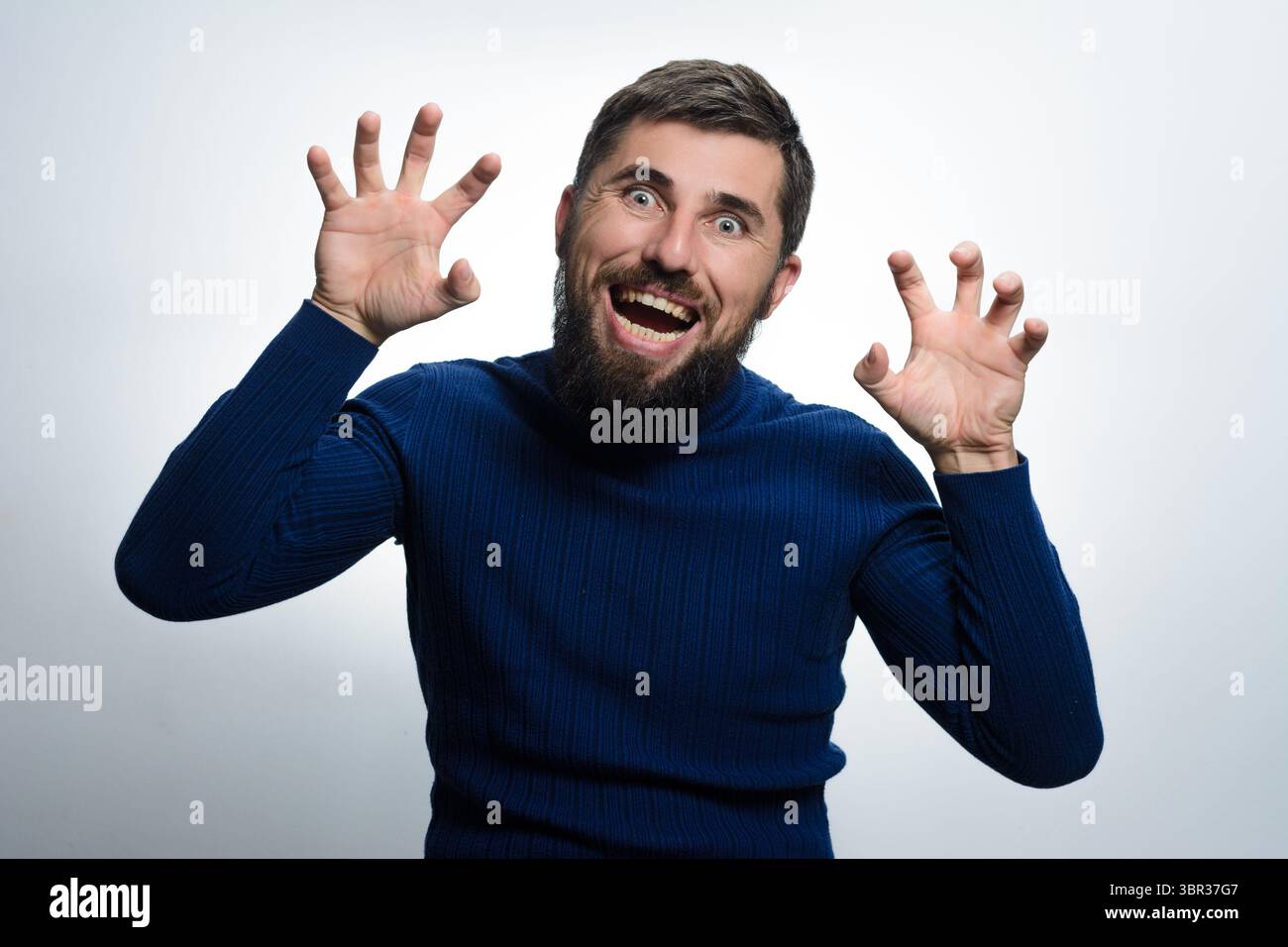 Ein bärtiger Mann in einem blauen Pullover bewegt sich spielerisch mit ausgestreckten Händen zur Kamera. Komischer Ausdruck auf seinem Gesicht gegen einen einfachen Hinterwäldler Stockfoto
