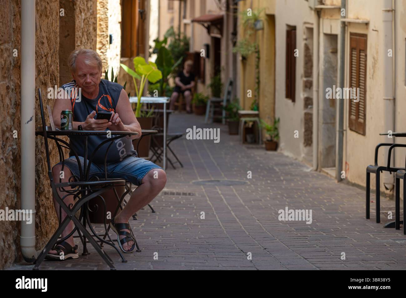 Chania, Kreta, Griechenland - 22. Juni 2025: Mann sitzt an einem Café-Tisch in einer engen Gasse von Chania, während er sein Telefon benutzt. Stockfoto