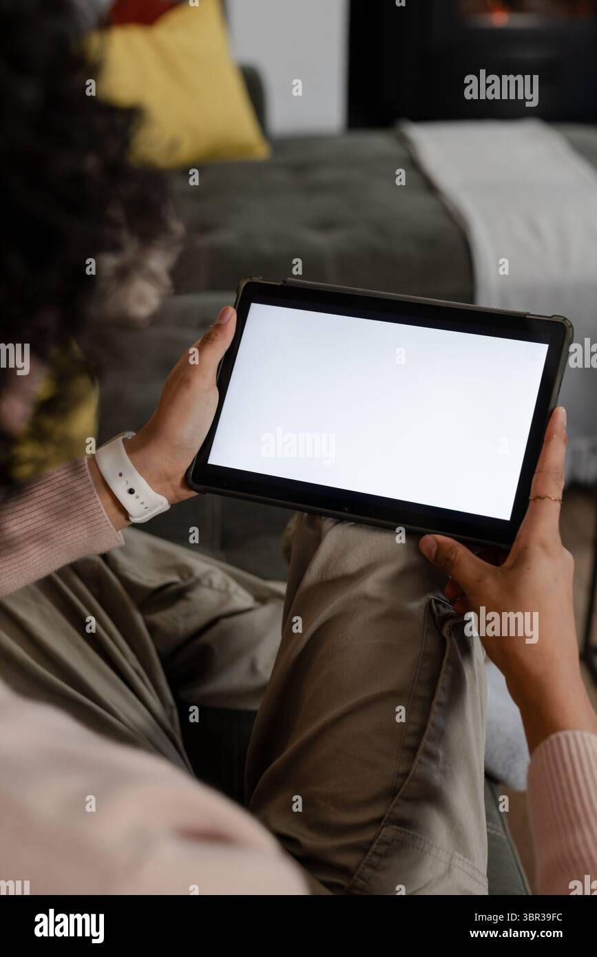 Nicht binäre Person, die auf olivgrünem Sofa sitzt und ein Tablet mit weißem Bildschirm im Wohnzimmer hält Stockfoto