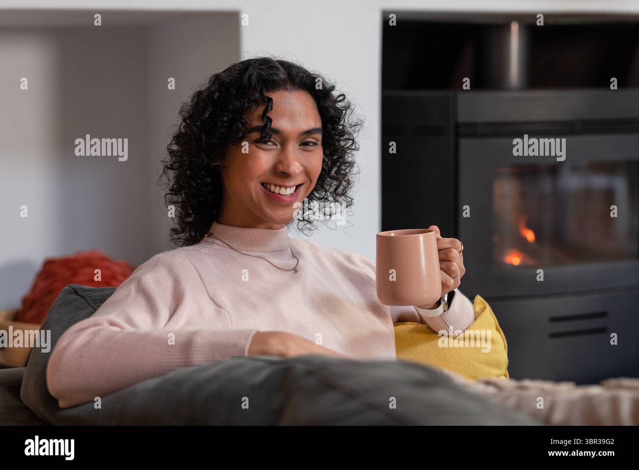 Nicht-binäre Person, die auf der Wohnzimmercouch sitzt und einen rosafarbenen Becher mit Herd hält, der warmes Licht gibt Stockfoto