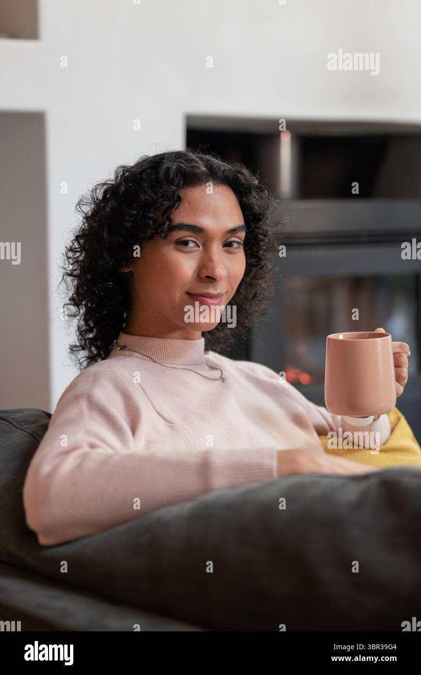 Nicht binäre Person mit rosa Becher, die auf grauem Sofa zu Hause mit Kamin und gelbem Kissen sitzt Stockfoto