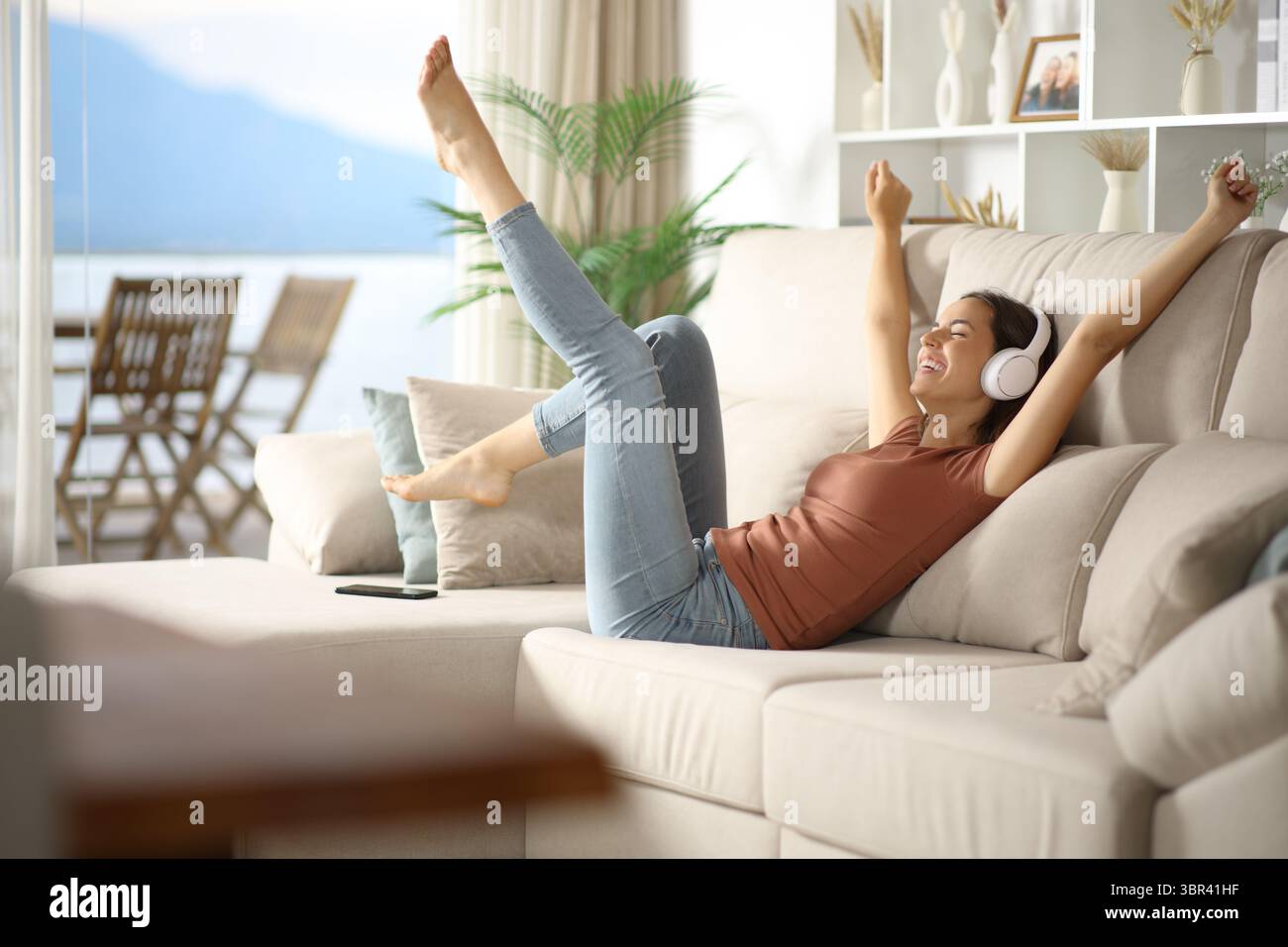 Aufgeregte Frau, die auf einem Sofa tanzt und zuhause Musik über Kopfhörer hört Stockfoto
