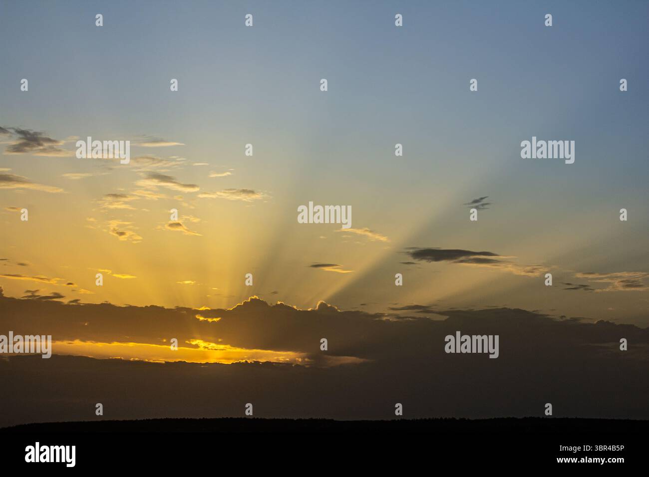 Der Himmel zeigt eine brillante Darstellung, während die Sonne untergeht und goldene Strahlen durch Wolken strömen und den Horizont in der Abenddämmerung erleuchten. Stockfoto