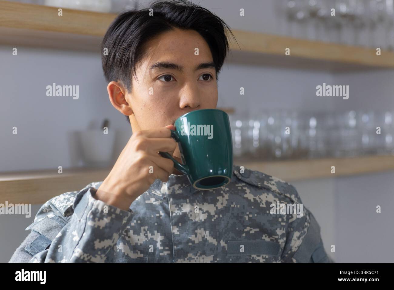 Asiatischer Mann in digitaler Camouflage-Uniform, der im militärischen Pausenbereich sitzt und an grünem Becher schlürft Stockfoto