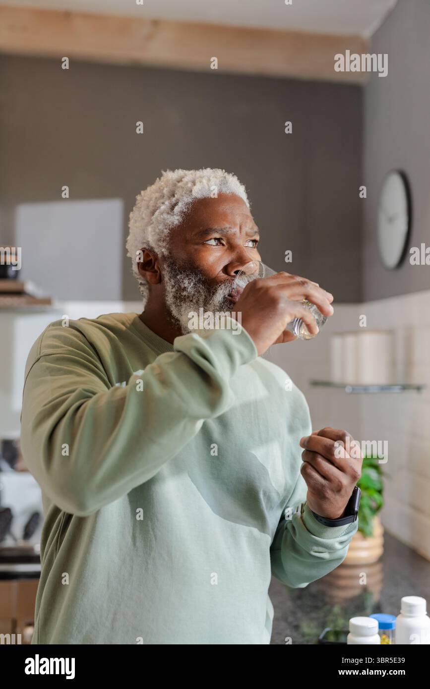Ranghoher Afroamerikaner trinkt Wasser aus klarem Glas in moderner Küche mit Gläsern auf Regalen Stockfoto
