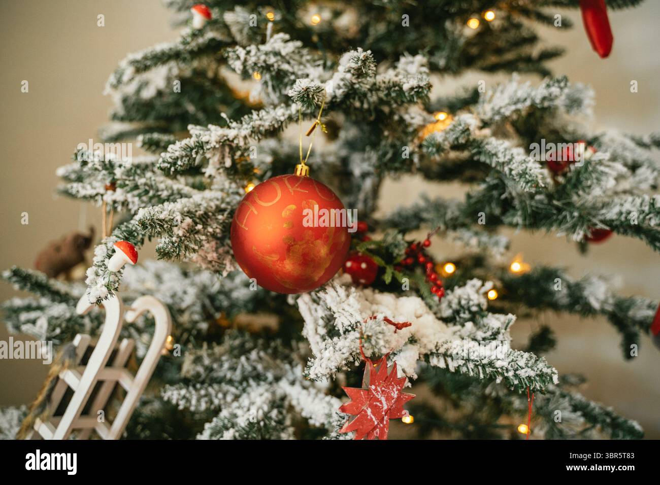 Schneebedeckter Weihnachtsbaum mit roten Ornamenten und festlichen Dekorationen, darunter ein Schlitten und handgefertigte Elemente. Warme Lichter sorgen für ein gemütliches h Stockfoto Schneebedeckter Weihnachtsbaum mit roten Ornamenten und festlichen Dekorationen, darunter ein Schlitten und handgefertigte Elemente. Warme Lichter sorgen für ein gemütliches h Stockfoto